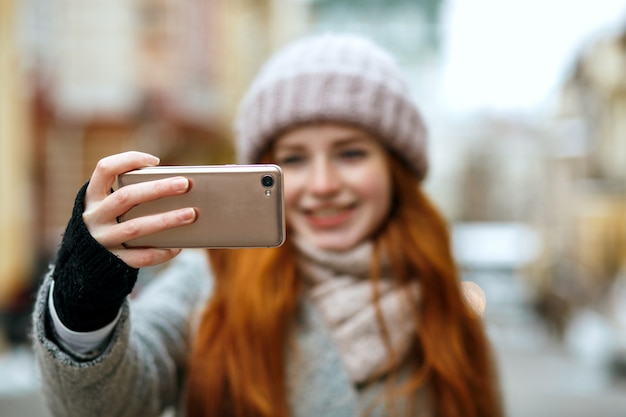 Joyeuse femme rousse en vêtements d'hiver prenant selfie sur son téléphone portable. Espace pour le texte
