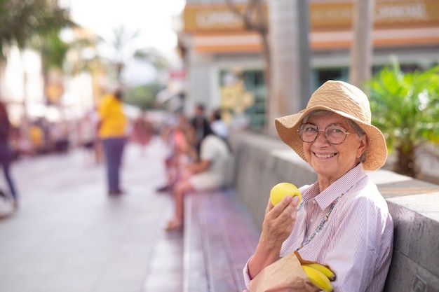 Joyeuse femme séduisante senior assise à l'extérieur dans la rue de la ville avec un sac en papier avec des pommes et des bananes tenant une pomme jaune concept de retraité détendu et d'une alimentation saine