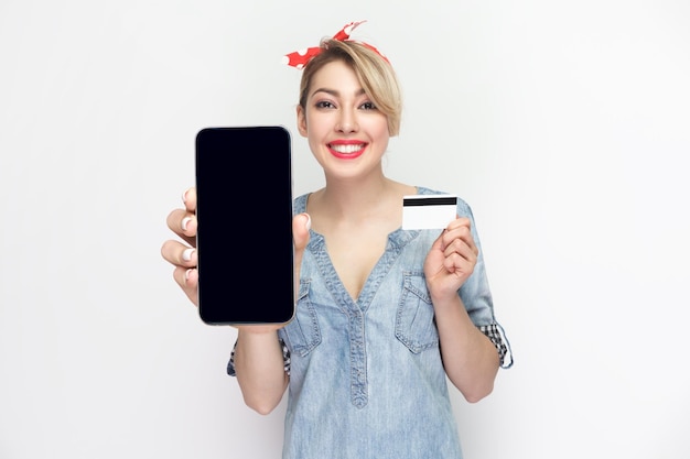 Joyeuse femme souriante en chemise en jean bleu montrant un téléphone avec un écran vide et une carte de crédit