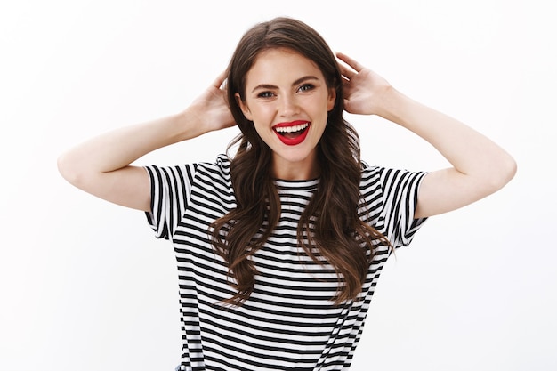 Joyeuse femme souriante, détendue et insouciante avec du rouge à lèvres rouge, un t-shirt rayé, se tenir les mains derrière la tête au repos, profiter des vacances d'été au bord de la piscine, s'amuser, mur blanc debout