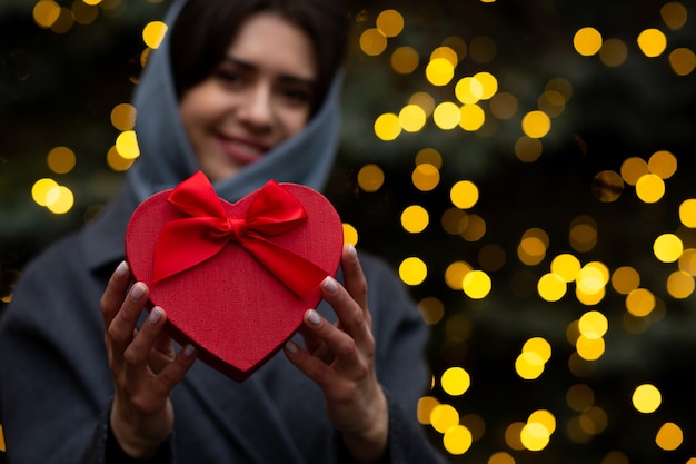 Joyeuse femme tenant une boîte-cadeau en forme de coeur pour la Saint-Valentin avec un arc sur un fond de lumières floues. Espace pour le texte