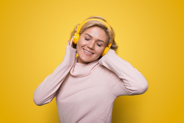 Joyeuse femme en vêtements décontractés dansant et écoutant de la musique avec plaisir via des écouteurs isolés sur fond jaune