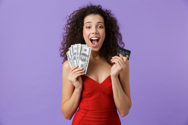 Joyeuse femme vêtue d'une robe rouge tenant un ventilateur d'argent et de carte de crédit, debout isolé sur mur violet