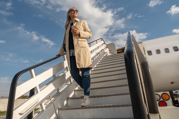 Joyeuse femme voyageuse en lunettes de soleil descendant dans les escaliers de l'avion après l'atterrissage à l'aéroport