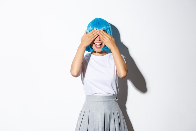 Joyeuse fêtarde en perruque bleue ferme les yeux avec les mains et souriant, en attente de surprise, debout.