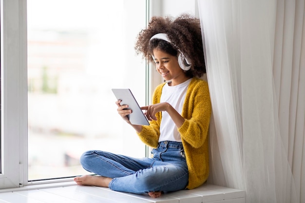 Joyeuse fille afro-américaine à l'aide d'une tablette numérique et d'un casque