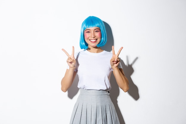 Joyeuse fille asiatique en perruque bleue, à la recherche de mignon en costume d'halloween et montrant des gestes de paix, debout kawaii.