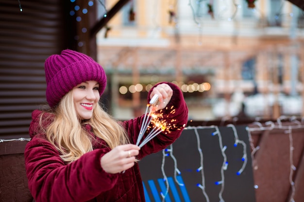 Joyeuse fille blonde vêtue de vêtements élégants, met le feu aux cierges magiques à la foire de Noël à Kiev