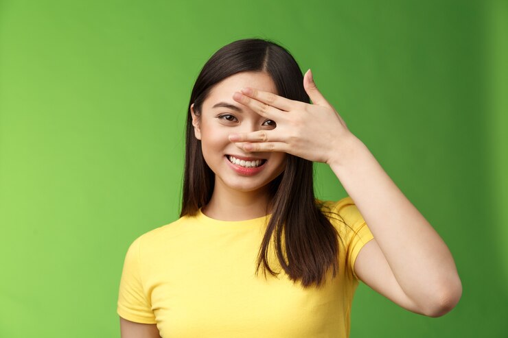 Photo joyeuse fille brune asiatique mignonne et enthousiaste souriant largement, sourire positif à pleines dents, yeux de couverture regardant à travers les doigts amusés, expression fascinée, fond vert debout