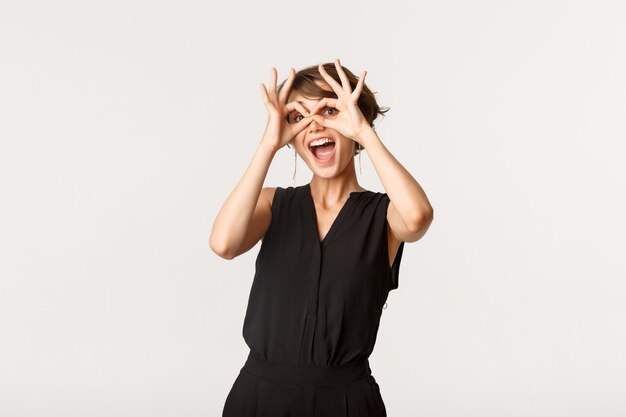 Joyeuse fille drôle montrant des lunettes à main et souriant étonné, debout blanc.