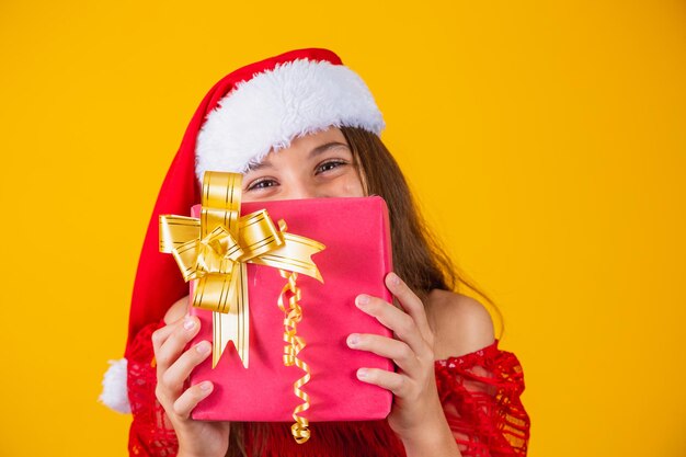 Joyeuse fille enfant drôle souriante en chapeau de père Noël rouge tenant un cadeau de Noël à la main et se cachant derrière un cadeau sur fond jaune.