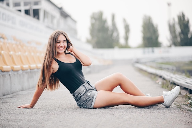 Joyeuse fille heureuse est assise sur la route sur fond de tribune Fille de bonne humeur à l'extérieur