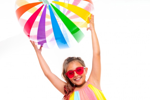 Joyeuse fille à lunettes de soleil tenant une boule rayée de couleur gonflable sur elle-même sur un mur blanc