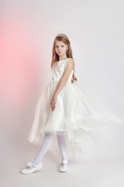 Joyeuse fille positive dans une belle robe de couleur blanche. Jeune fille s'amusant et posant sur fond gris. Émotions d'enfants lumineux