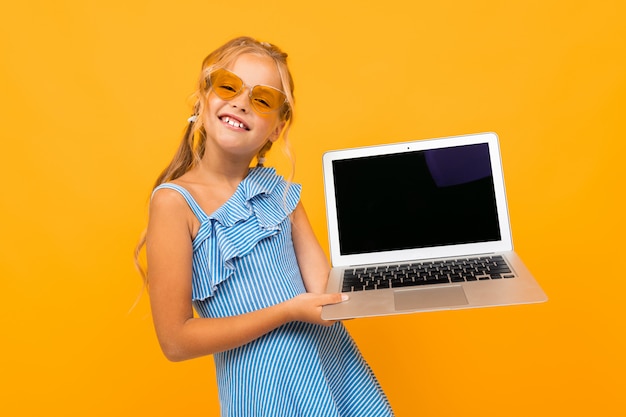 Joyeuse fille en robe sourit avec son ordinateur portable isolé sur fond jaune et orange