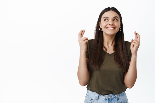 Joyeuse fille souriante ayant de l'espoir, croisant les doigts pour la bonne chance, faisant son vœu, veut quelque chose et en rêve, debout contre un mur blanc