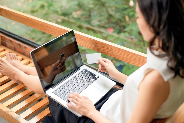 Joyeuse fille tenant une carte de crédit et utilisant un ordinateur portable assis sur un canapé à la maison