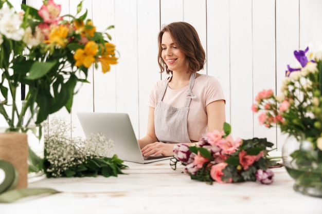 Joyeuse fleuriste dame debout avec des fleurs à l'aide d'un ordinateur portable.