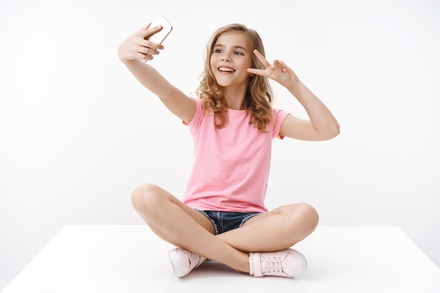 Joyeuse heureuse belle adolescente assise les jambes croisées sur le sol, jouant avec un smartphone, montre la paix, signe de la victoire sur un téléphone portable à l'avant, prenant un selfie, souriant optimiste, énergisé, mur blanc