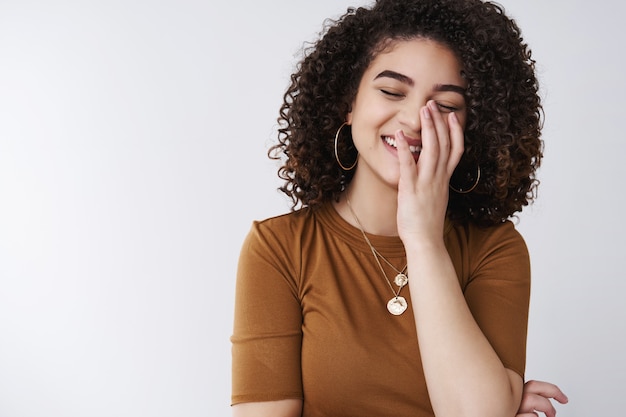 Joyeuse insouciante jeune fille élégante aux cheveux bouclés riant sincèrement les yeux fermés couvrent les dents blanches paume riant blague drôle debout mur blanc joyeux