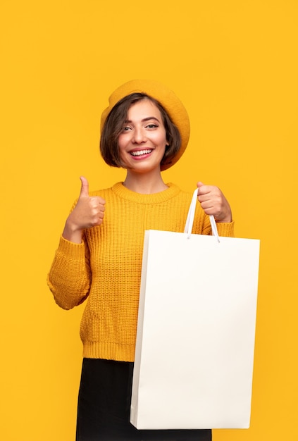 Joyeuse jeune femme accro du shopping dans des vêtements élégants montrant un sac en papier vierge et gesticulant le pouce sur fond jaune