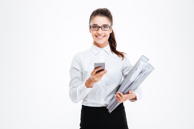 Joyeuse jeune femme d'affaires séduisante dans des verres tenant des dossiers et utilisant un téléphone portable sur un mur blanc