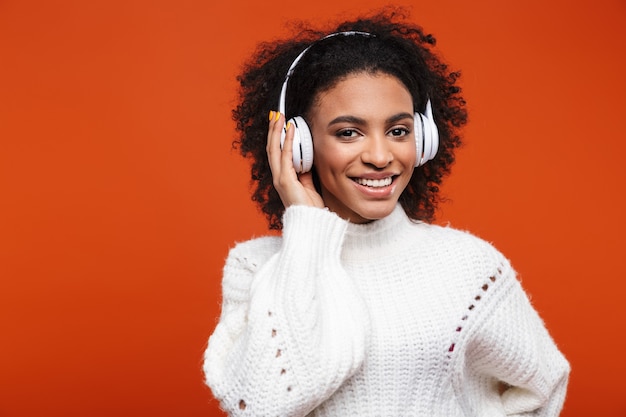 Joyeuse jeune femme africaine écoutant de la musique avec des écouteurs sans fil isolés sur un mur rouge