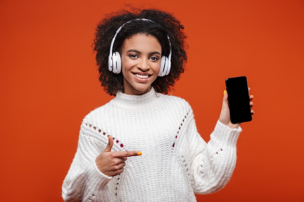 Joyeuse jeune femme africaine séduisante portant des écouteurs sans fil debout isolée sur un mur rouge, montrant un téléphone portable à écran blanc