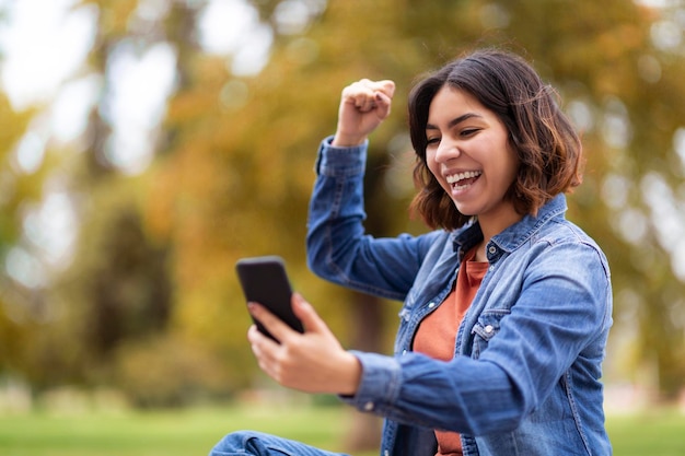 Joyeuse jeune femme arabe excitée célébrant le succès avec un smartphone à l'extérieur