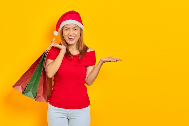 Joyeuse jeune femme asiatique en chapeau de père Noël tenant des sacs à provisions et montrant un espace de copie dans les paumes