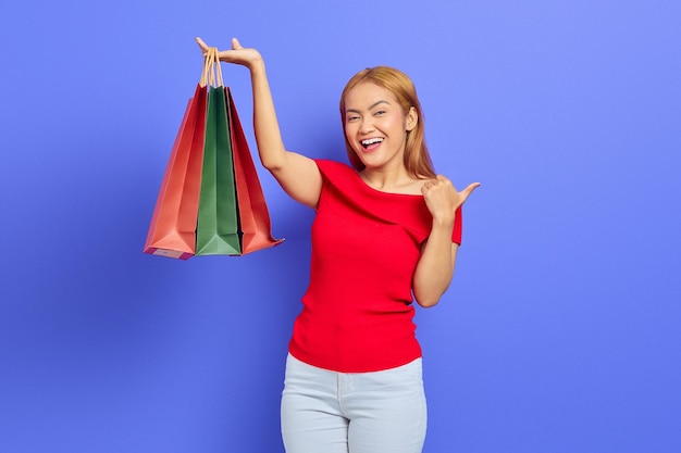 Joyeuse jeune femme asiatique montrant des sacs à provisions et pointant du doigt l'espace de copie isolé sur fond violet