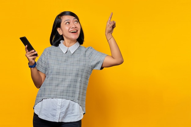Joyeuse jeune femme asiatique tenant un téléphone portable et pointant le doigt vers le haut sur fond jaune