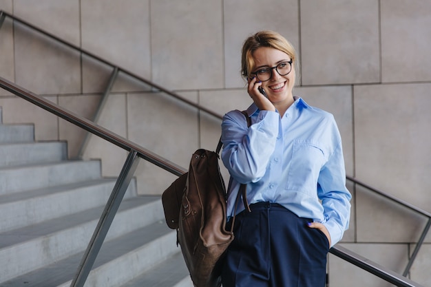 Joyeuse jeune femme aux cheveux blonds debout dans la rue et parlant sur mobile. Femme heureuse à lunettes utilisant un gadget moderne pour la conversation à l'extérieur.