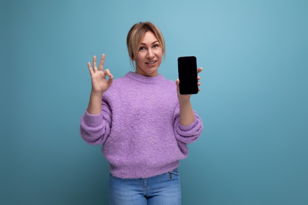 Joyeuse jeune femme blonde consultante en pull violet montre une maquette de smartphone sur fond bleu