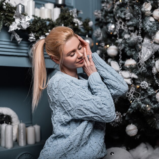 Joyeuse jeune femme blonde glamour dans un pull tricoté à la mode se détend dans un salon confortable près d'un arbre de Noël et d'une cheminée vintage