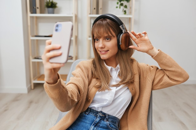 Joyeuse jeune femme blonde en pull chaud dans les écouteurs écouter des chansons préférées faisant un appel vidéo selfie assis dans un fauteuil à l'intérieur de la maison moderne Temps de musique Relaxant Cool playlist Concept Copier l'espace