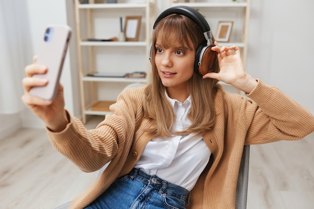 Joyeuse jeune femme blonde en pull chaud dans les écouteurs écouter des chansons préférées faisant un appel vidéo selfie assis dans un fauteuil à l'intérieur de la maison moderne Temps de musique Relaxant Cool playlist Concept Copier l'espace