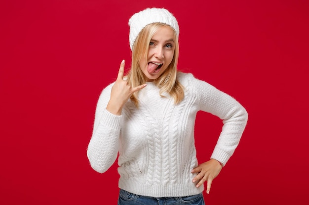 Photo joyeuse jeune femme en chapeau pull blanc isolé sur fond de mur rouge en studio. concept de saison froide de mode de vie sain. maquette de l'espace de copie. montrant la langue, représentant un signe de rock en métal lourd.