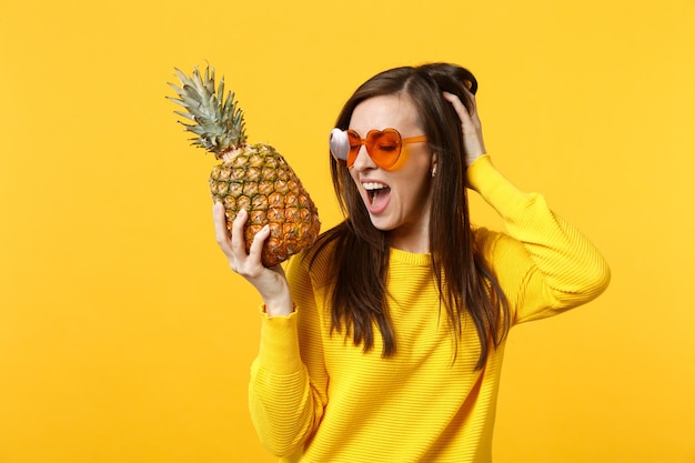 Joyeuse jeune femme dans des lunettes de coeur mettant la main sur la tête tenant des fruits frais d'ananas mûrs isolés sur fond orange jaune. Mode de vie vivant des gens, concept de vacances relaxantes. Maquette d'espace de copie
