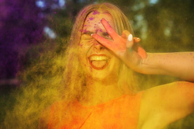 Joyeuse jeune femme debout dans un nuage de peinture jaune Holi montrant un geste de paix