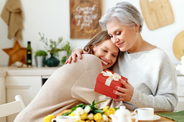 Joyeuse jeune femme étreignant une femme âgée heureuse avec les yeux fermés et donnant des fleurs et une boîte cadeau le jour de la fête des mères