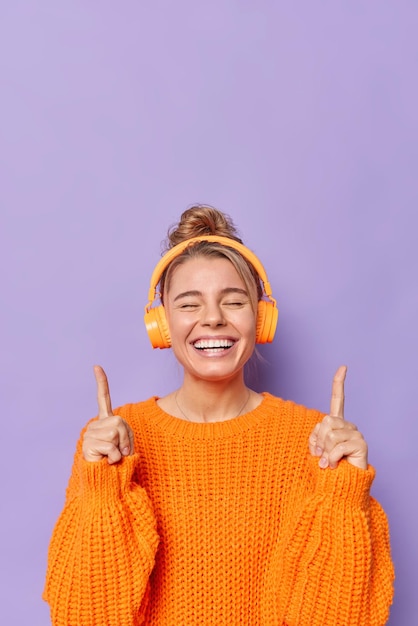 Joyeuse jeune femme européenne garde les yeux fermés sourit largement les deux index au-dessus porte un pull orange tricoté écoute de la musique via des écouteurs stéréo isolés sur fond violet.