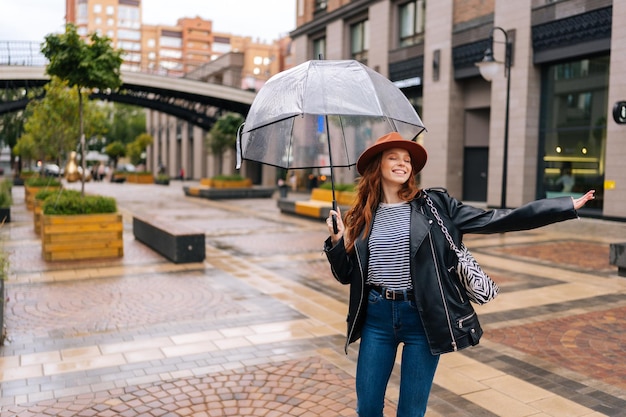 Joyeuse jeune femme excitée au chapeau élégant dansant et s'amusant avec un parapluie transparent sur la belle rue de la ville profitant du temps pluvieux à l'extérieur