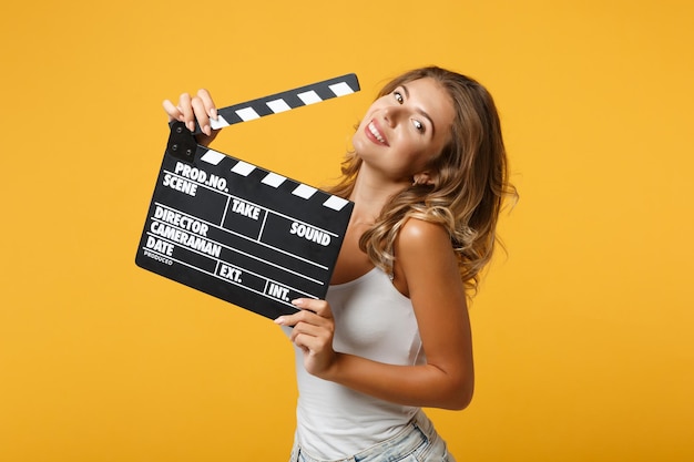 Joyeuse jeune femme fille dans des vêtements décontractés légers posant isolé sur fond orange jaune en studio. Concept de style de vie des gens. Maquette de l'espace de copie. Tenant un clap de cinéma noir classique.