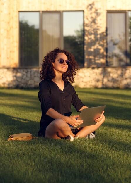 Joyeuse jeune femme à lunettes de soleil assise sur l'herbe avec les jambes croisées pendant la journée tout en utilisant un ordinateur portable et en discutant