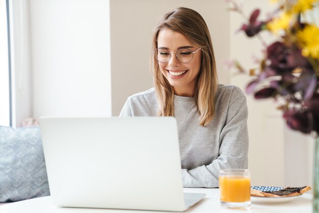 joyeuse jeune femme à lunettes utilisant un ordinateur portable tout en prenant son petit-déjeuner dans le salon