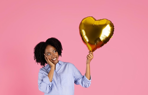 Joyeuse jeune femme noire en tenue décontractée regarde un ballon gonflable en forme de cœur doré