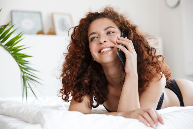 Joyeuse jeune femme optimiste positive et positive en lingerie à l'intérieur de l'hôtel à domicile sur le lit en parlant par téléphone portable.