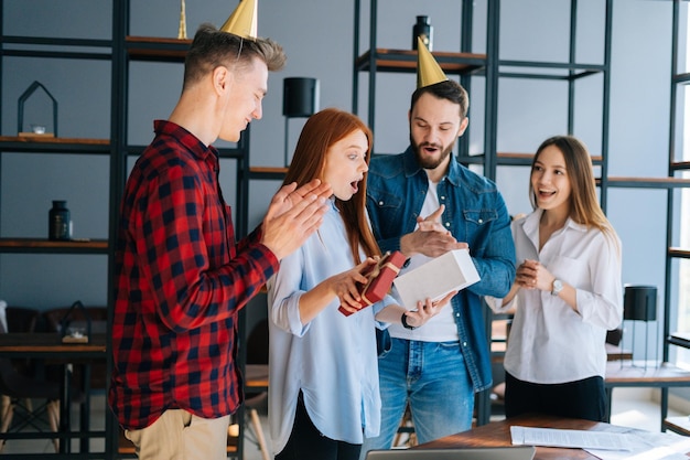 Joyeuse jeune femme ouvrant le cadeau offert par des collègues de bureau aux applaudissements de ses collègues