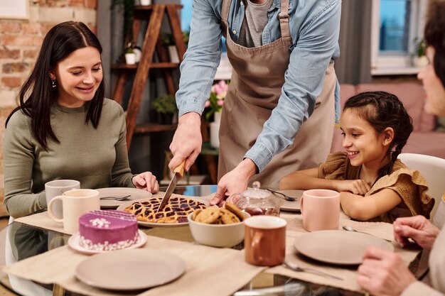 Joyeuse jeune femme et petite fille assise près de la table servie et regardant l'homme en tablier coupant une tarte maison sucrée sur une assiette avant de prendre le thé